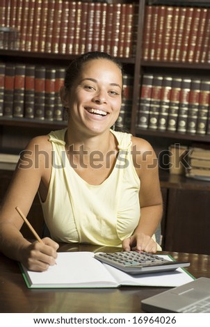 Happy student writing in a notebook with a pencil, and a laptop on her desk and books in the background. Vertically framed photo.