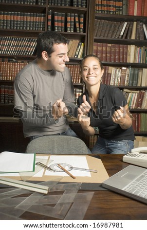 Two students joking around in library with paperwork, drafting tools, phone, and rulers on desk. Vertically framed photo.