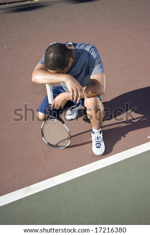 Tennis player crouching down looking defeated and sad, he holds his tennis racket and hangs his head in shame. Vertically framed photo.