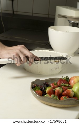 Hands using a knife to smooth out the icing on a cake. There is a fruit plate next to it. Vertically framed photo.