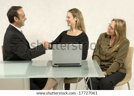 Two women and a man in a meeting at a desk in front of a computer. The man and a woman are shaking hands. Horizontally framed photo.