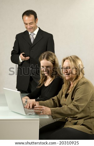 Two women sitting at a desk one is typing on a computer, the other is watching and a man is standing looking at his phone. They are all smiling. Vertically framed photo.