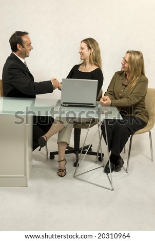 Two women and a man seated at a desk in front of a laptop, they are all smiling and the man and one woman are shaking hands. Vertically framed photo.