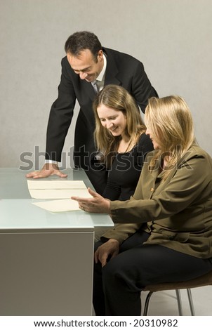 Three people are in a business meeting.  They are smiling and looking at some pieces of paper on the table.  The women are sitting and the man is standing.  Vertically framed shot.