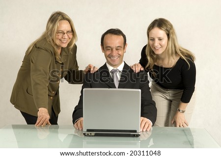 Two women standing over a man seated at a desk with their hands upon his shoulders as they all smile. Horizontally framed photo.