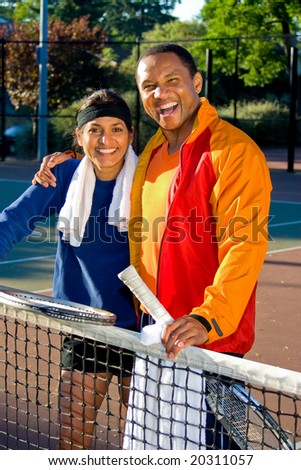 Man and woman tennis players laughing as they stand at the net. He has his arm around her and they are holding rackets. Vertically framed photo.