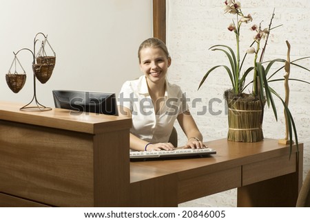 Smiling young woman sitting at a desk talking on the phone. There is a computer and a plant on the desk. Horizontally framed photo.