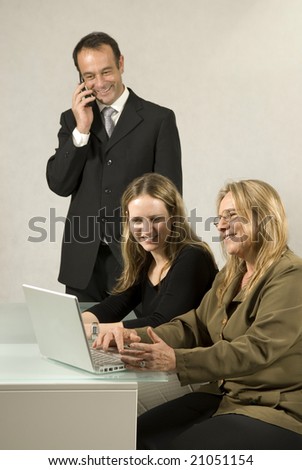 Two women at a desk and a man standing talking on his cell phone. All are smiling and looking at paperwork.