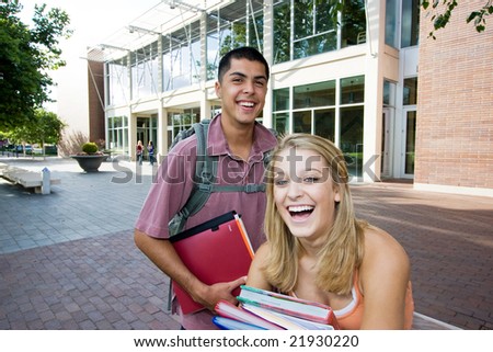 Two students one girl and one boy carrying their books outside of school. They are laughing. Horizontally framed photo.