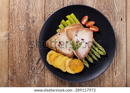 Grilled tuna steak served on asparagus with roasted potatoes on a black plate.