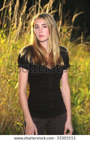 Pretty blond teenager in black blouse standing in front of tall grass facing straight at camera