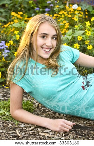 Pretty smiling blond girl in light blue blouse resting on elbow lying in front of flowers.