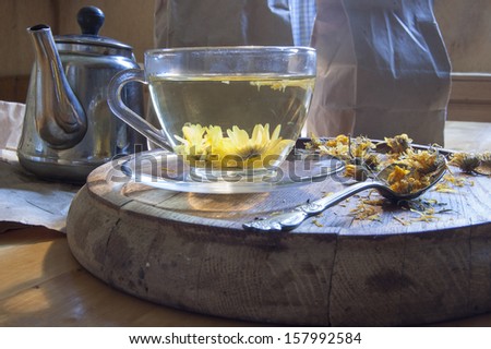 A cup of herbal tea. Near kettle, tea spoon and dried marigold flowers on a wooden board.