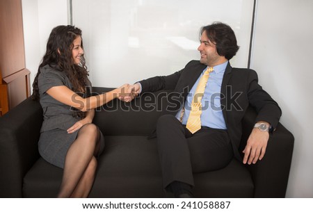 Two business persons are sitting and shaking hands on black sofa at office