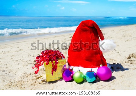 Christmas hat with gift box and christmas balls on the beach by the ocean - holiday concept