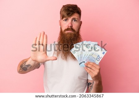 Young caucasian ginger man with long beard holding banknotes isolated on pink background standing with outstretched hand showing stop sign, preventing you. Foto stock © 