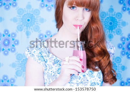 Vintage fashion styled redheaded woman drinking through a straw from a bottle of pink soda, wearing a blue patterned dress against a blue patterned backdrop (urban camouflage)