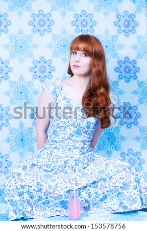 Vintage fashion styled red headed woman sitting cross legged with arms behind her with a coy expression, wearing a blue patterned dress against a blue patterned backdrop (urban camouflage)