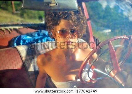 Attractive young lady looking through her sunglasses through the windshield of an older car with a red interior.