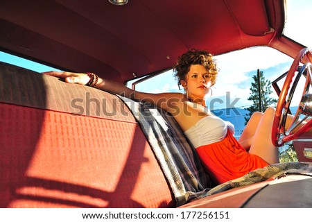 Attractive, sultry woman on a sunny day wearing an orange and white dress sitting in the drivers seat of an old red car with bench seats.