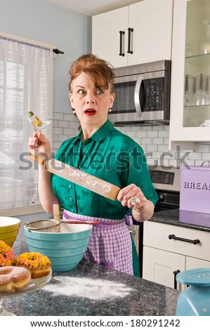 An attractive middle-aged woman in a kitchen wearing a vintage dress, holding a martini in one hand and a rolling pin in the other with cooking items around her, looking off camera with fear