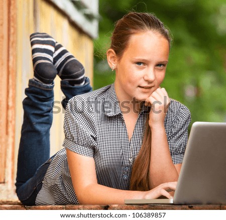 Young girl (looking at the camera) lying on the porch of the rural house with a laptop.