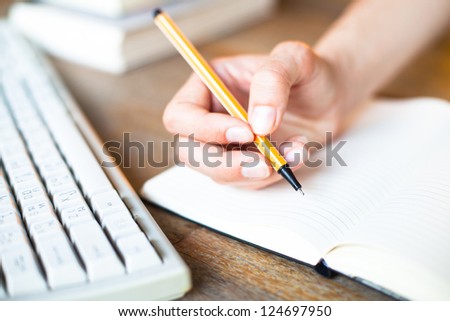 Hands writes a pen in a notebook (computer keyboard, a stack of books in background)