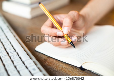 Hands writes a pen in a notebook, computer keyboard and a stack of books in background.