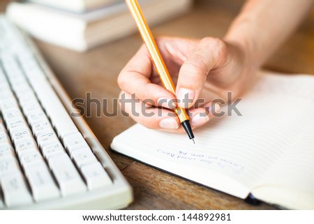 Hands writes a pen in a notebook, computer keyboard in background.