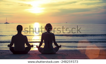 Silhouette of young man and woman practicing yoga in the lotus position on the ocean beach during amazing sunset.