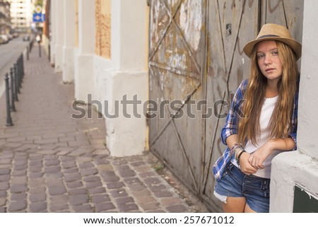 Young cute long haired girl standing on a street corner in shorts and hat.