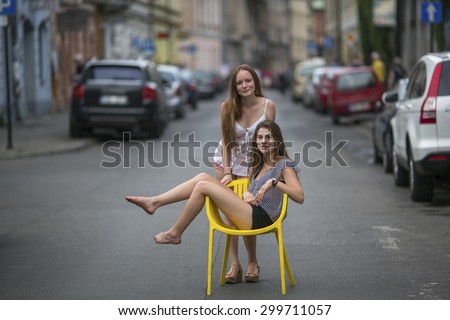 Two young girls girlfriend. Conceptual photo, a teen girl sits on a chair in the middle of the street, her friend stands behind her. European city blur in the background.