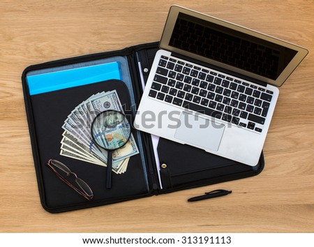 US hundred dollar bills under magnifying glass on business folder and Laptop lying on oak wood texture, top view.