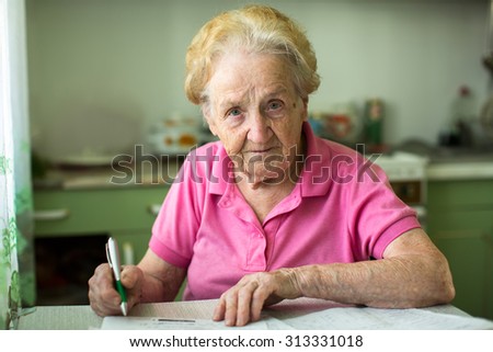 Elderly senior woman populates handle her utility bills notices, sitting at the table in the kitchen.