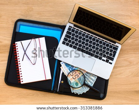 Laptop, notebook, glasses and banknotes of euros and dollars laid out on the business folder lying on the wood texture.
