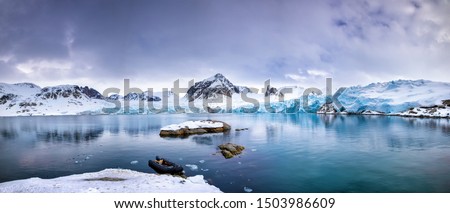 Panarama of the mountains, snow and blue glacial ice of the Smeerenburg glacier, Svalbard, and archipelago between mainland Norway and the North Pole. An inflatable boat is anchored in the foreground,