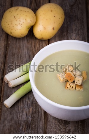 A bowl of leek and potato soup with bread croutons, over old wood table with fresh leeks and potatoes alongside