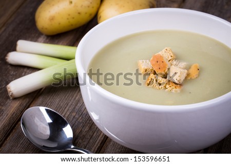 A bowl of leek and potato soup with bread croutons, over old wood table with fresh leeks and potatoes alongside