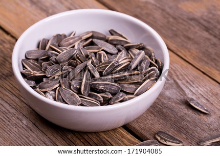 Roasted and salted sunflowers seeds in their shells, in white ceramic bowl over old wood background.