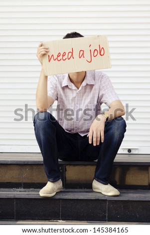 Unemployed young man sitting on the steps of a closed shop window with a cardboard sign