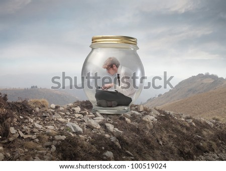 Young businessman trapped in a glass jar and using a laptop on a mountain peak