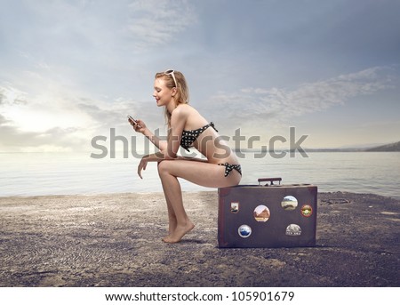 Beautiful young woman in bikini sitting on a suitcase and using a mobile phone