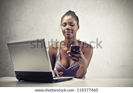 A beautiful black woman is smiling while is using a smart-phone and a laptop computer