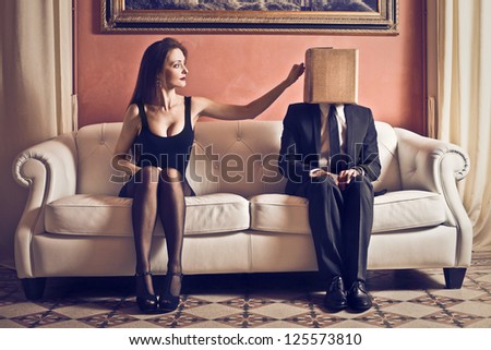 Elegant redhead woman sitting on a white sofa touching a box that covers the face of a businessman on the sofa