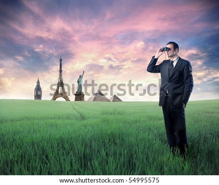 Businessman standing on a green meadow and using binoculars with landmarks from different cities on the background