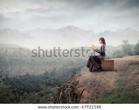Woman sitting on a suitcase and reading a book with landscape on the background