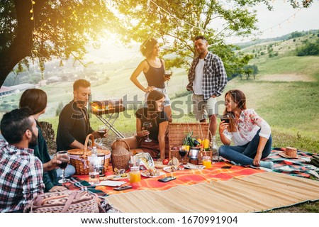 Picnic in the countryside. Group of young friends, at sunset on spring day are sitting on the ground in a park near trees. They drinking red wine and eating grilled meat with barbecue