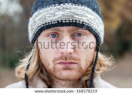 Portrait, twenty four year old man with blue eyes and long blonde hair, wearing a woolen hat in a day of winter in a forest.