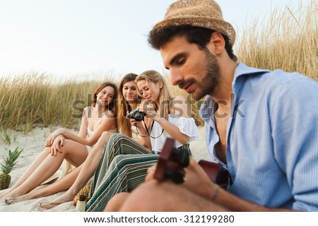 Beach at sunset. A group of young friends (a man and three women) are on the beach to relax after a day at sea, the man plays guitar while the three beautiful women taking pictures and observe