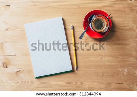A recycled paper notebook checked with a black pencil with the eraser at the top and a cup of coffee with red saucer and spoon, are arranged on a brown wooden table. View from the top
